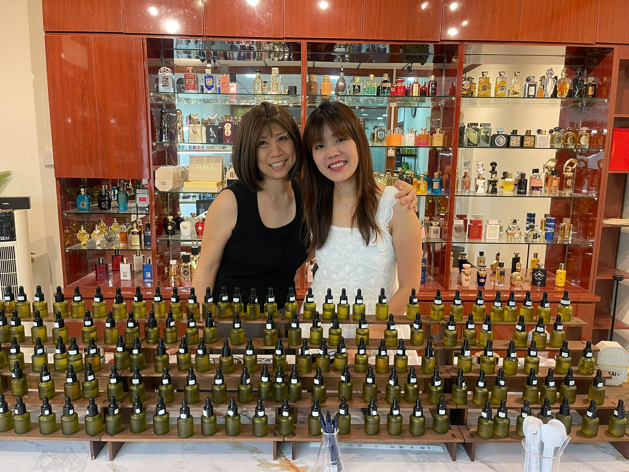 Two women standing behind a display of small bottles, with shelves of various containers in the background.