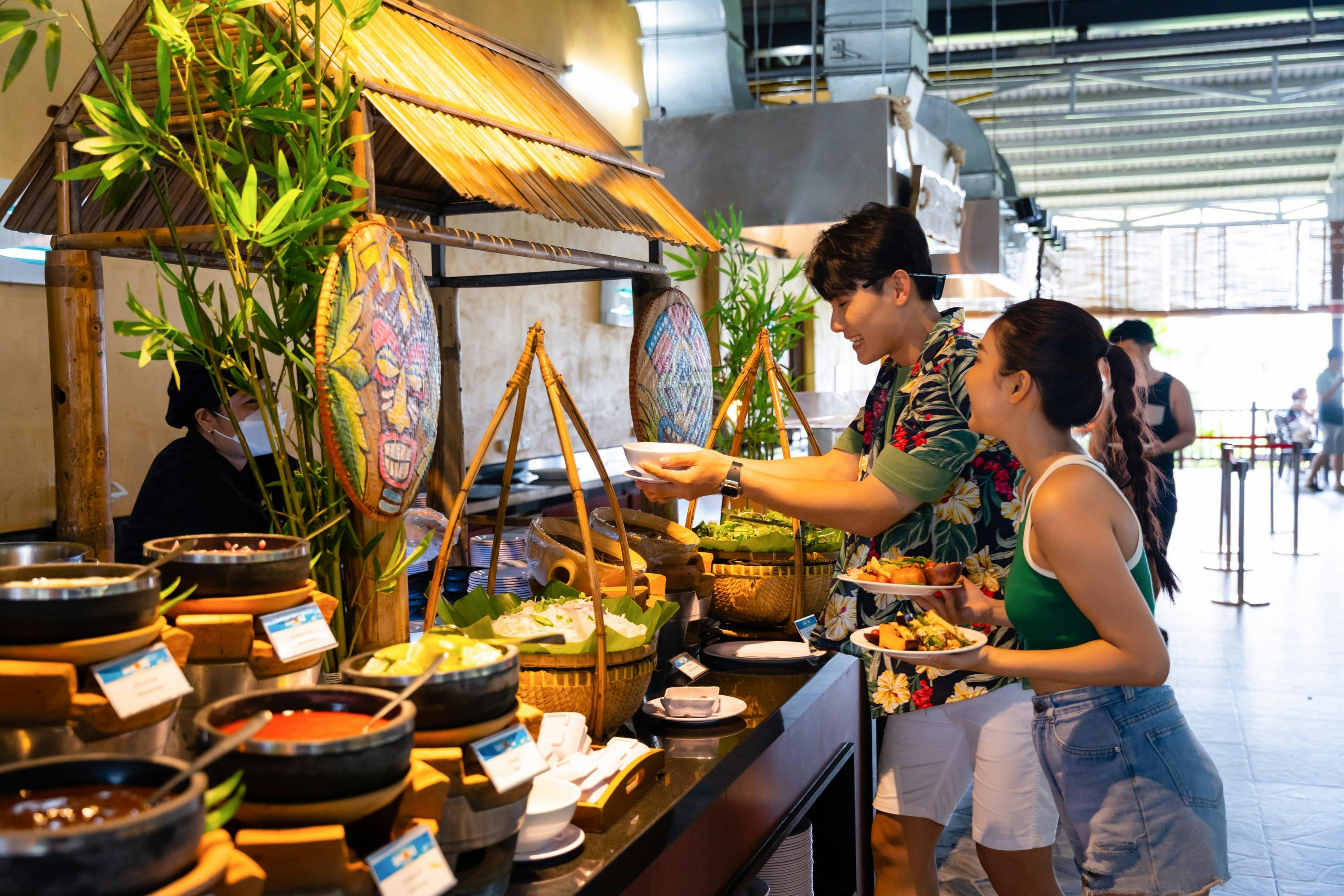 Two people serving food from a buffet in a well-lit restaurant, surrounded by various decorative elements and food baskets.