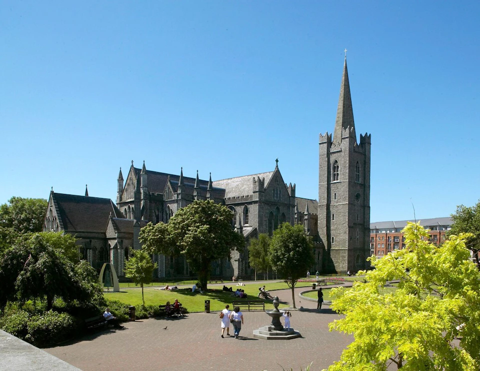 A Gothic-style cathedral with a tall spire, surrounded by trees and people walking in a sunlit courtyard on a clear day.