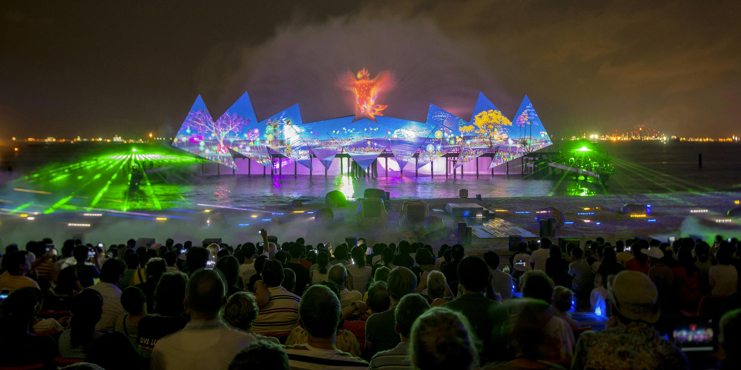 Dav sleduje živou, osvětlenou vodní a světelnou show s barevnými projekcemi a noční mlhou.