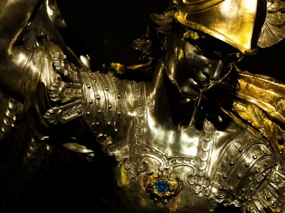 Close-up of a metallic statue depicting a warrior in detailed armor with a blue gemstone and winged shoulder pads.