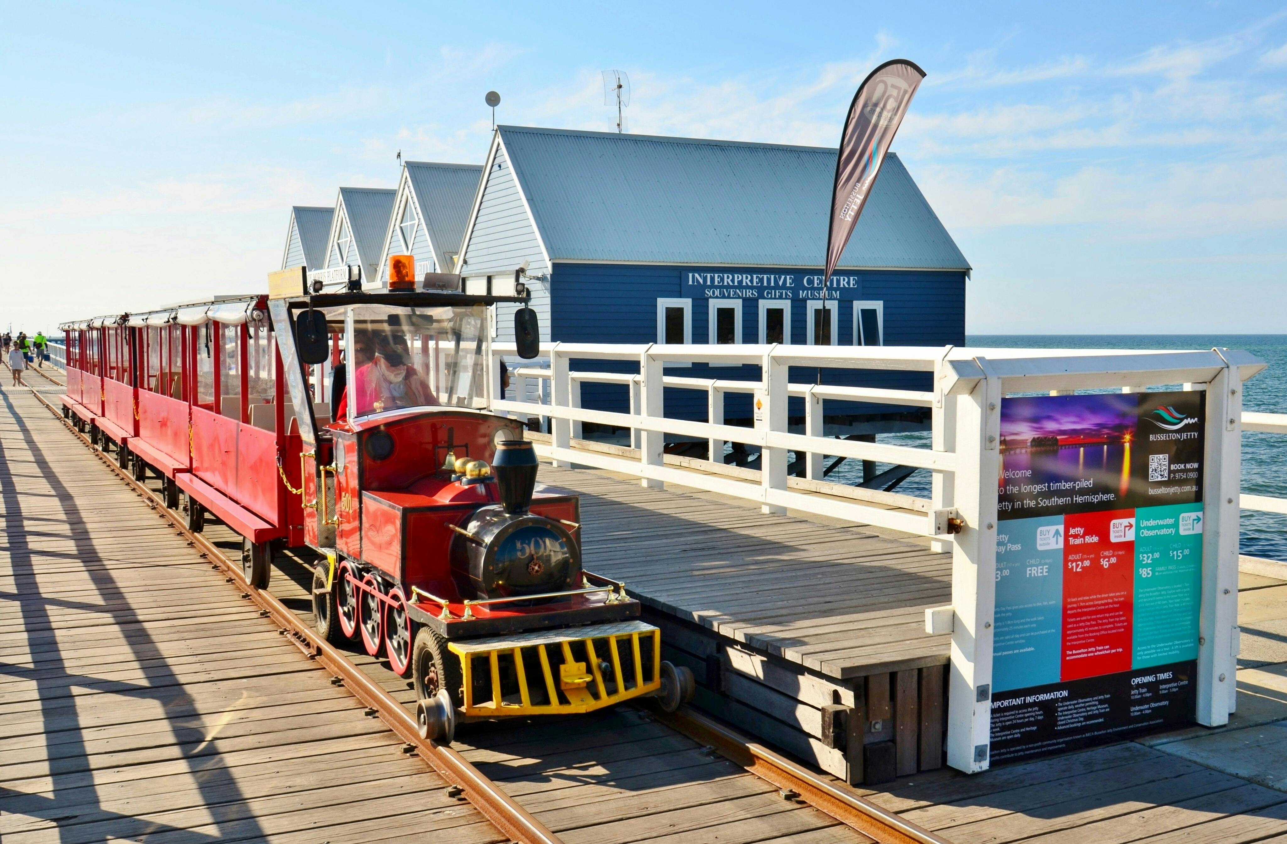 Un train touristique rouge sur rails circule le long d'une jetée en bois, à côté d'un bâtiment bleu portant l'inscription "Centre d'interprétation".