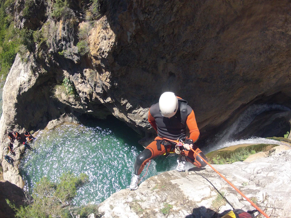 Person in orange and black wetsuit and helmet rappelling down a cliffside above a green water pool.