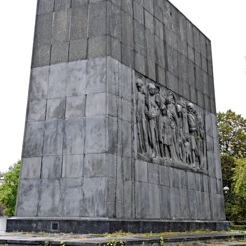 Ett högt, grått monument med en basrelief som visar en grupp mänskliga figurer. Omgivande trappsteg har gräsfläckar.