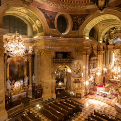 Utsmyckad kyrkinteriör med höga valv, detaljerade fresker, statyer, förgyllda accenter och dekorerad predikstol. Bänkrader i förgrunden.