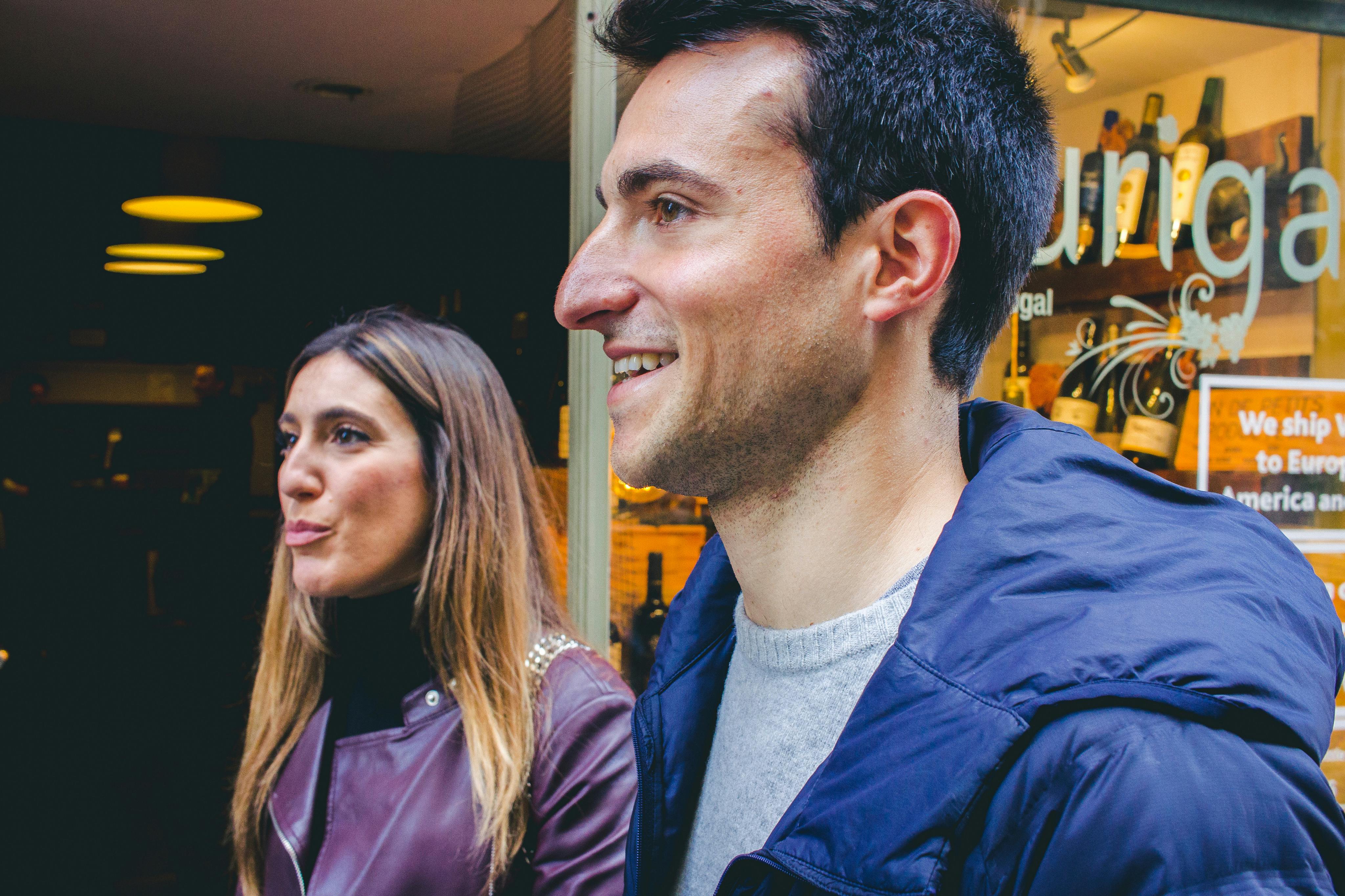 Een man en vrouw staan samen en glimlachen in een binnenhuis met warme verlichting op de achtergrond.