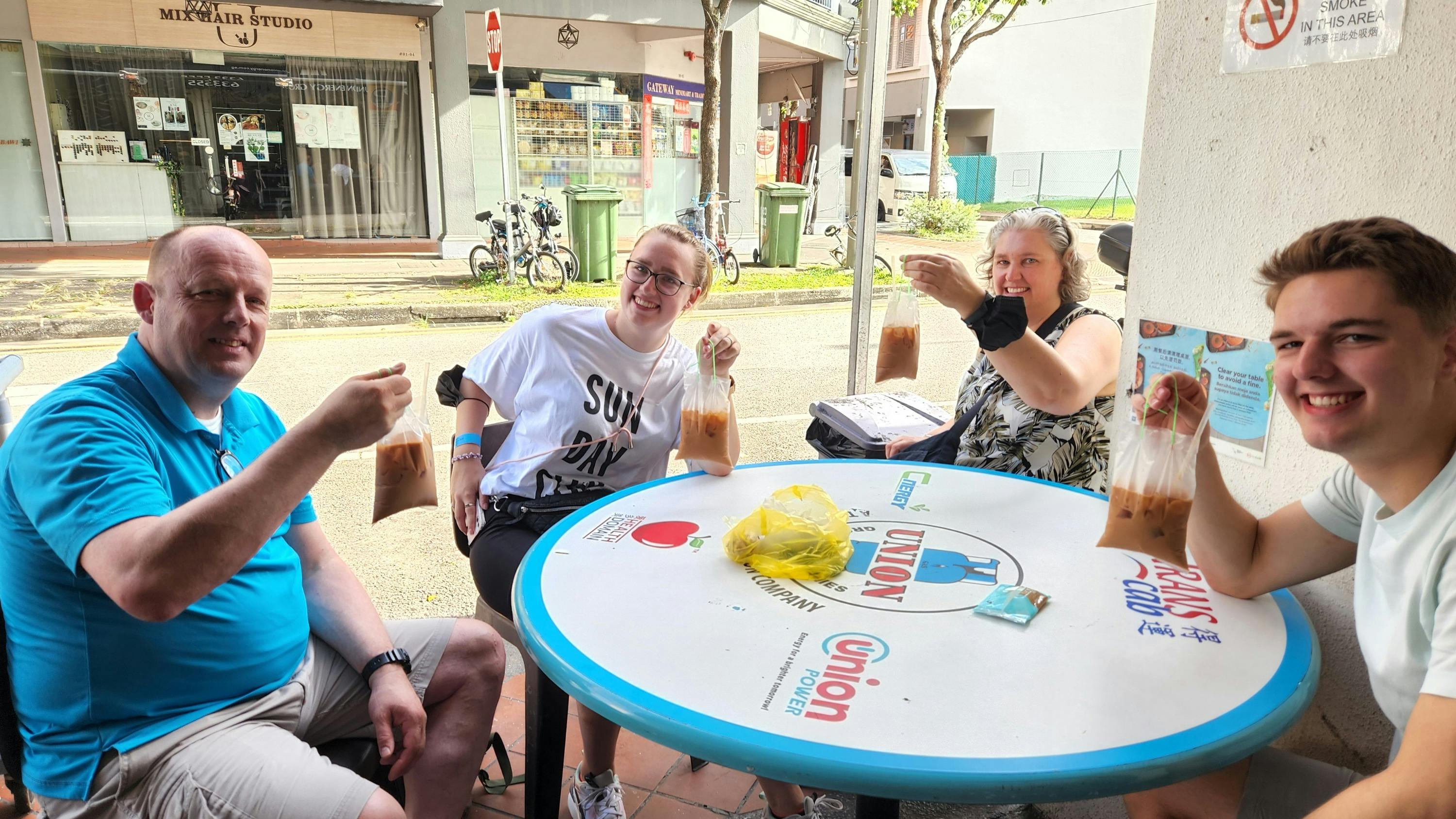 Three people sitting around a table outdoors, smiling and holding clear bags with a brown liquid, likely beverages.