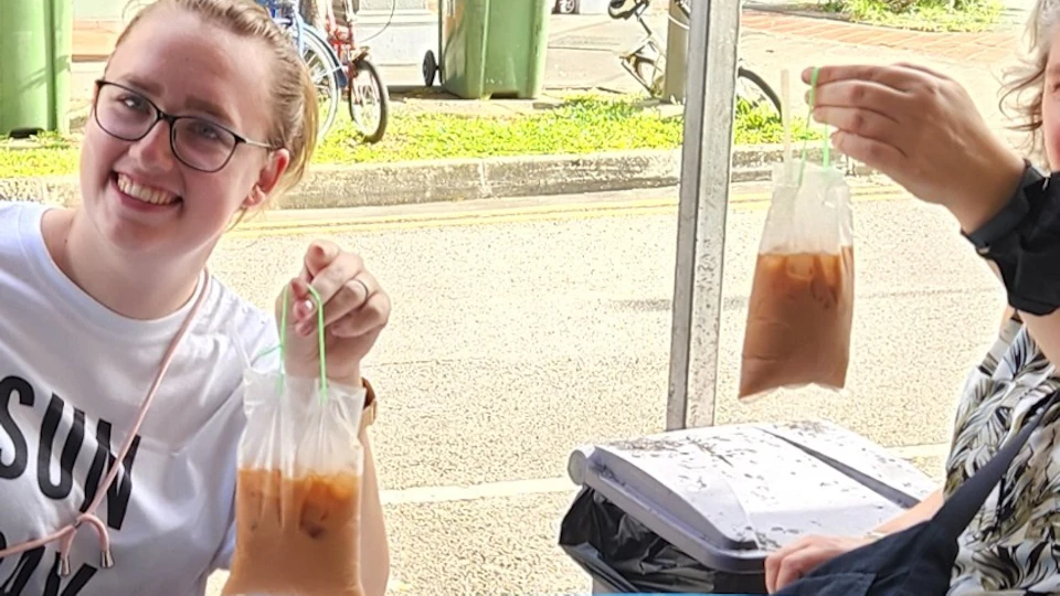Three people sitting around a table outdoors, smiling and holding clear bags with a brown liquid, likely beverages.