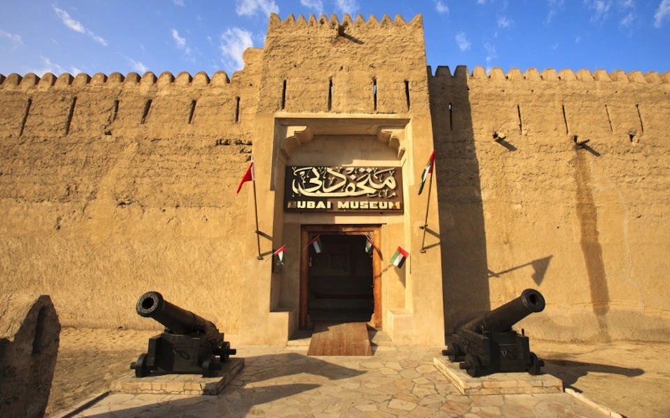 Entrance of Dubai Museum with old cannons on either side, UAE flags, and a sandy-colored stone facade under a blue sky.