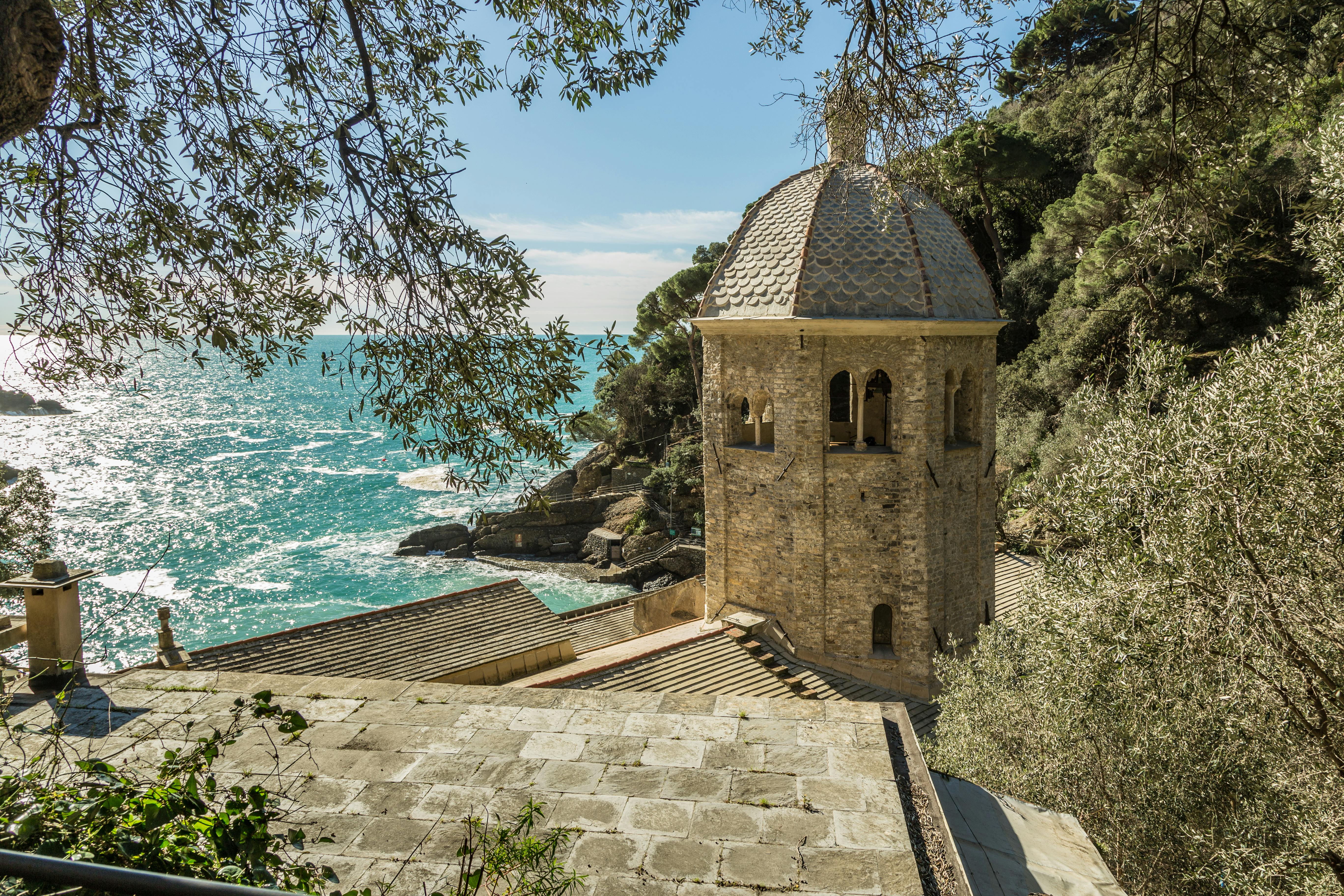 石头建筑，穹顶可俯瞰岩石海岸线和绿松石海，四周环绕着树木和树叶。