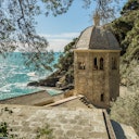 Stone building with a dome roof overlooking a rocky coastline and turquoise sea, surrounded by trees and foliage.