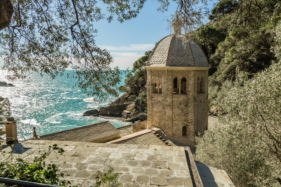 Stone building with a dome roof overlooking a rocky coastline and turquoise sea, surrounded by trees and foliage.