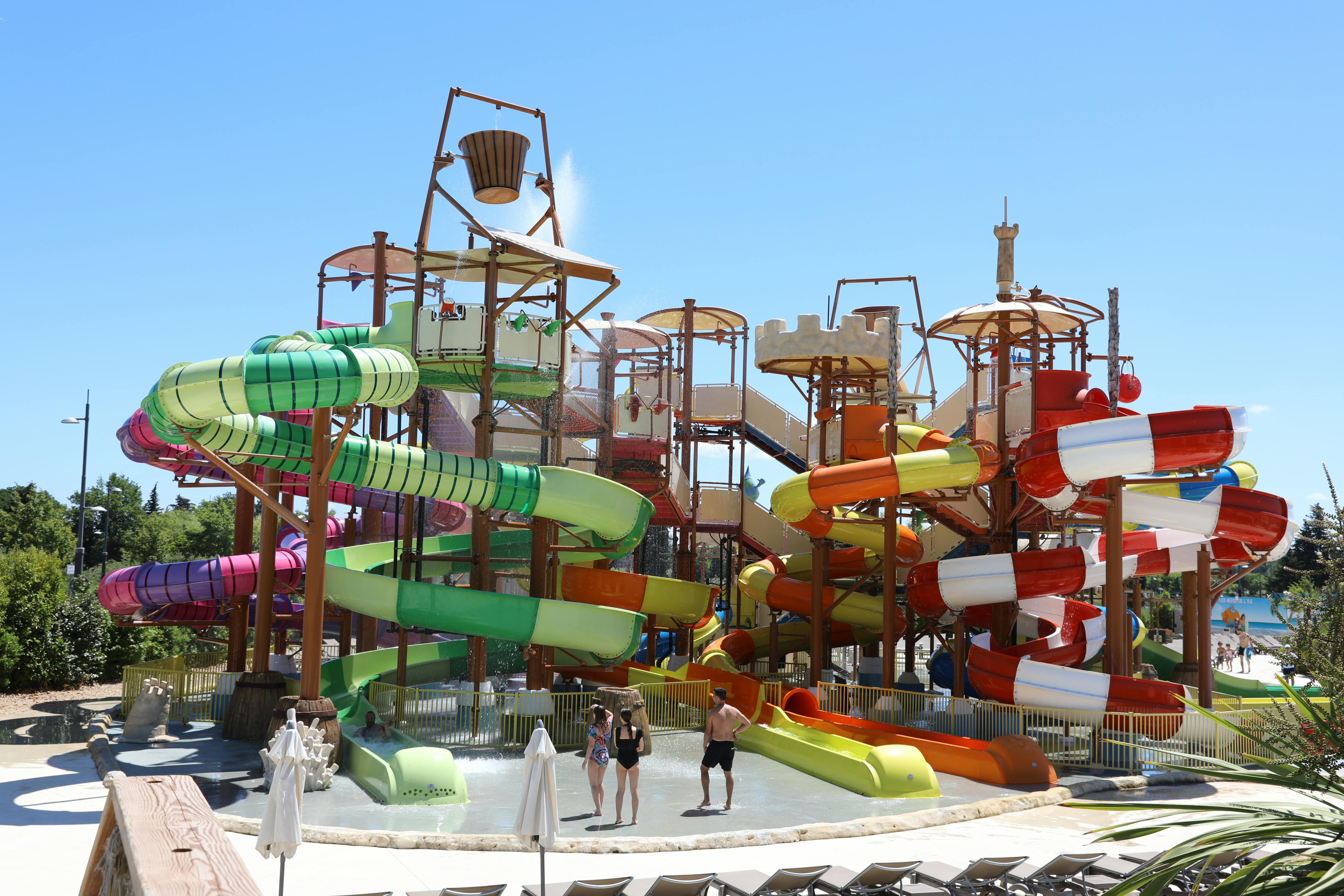 A colorful water playground with multiple slides and a large tipping bucket. Three people stand at the base near small pools.