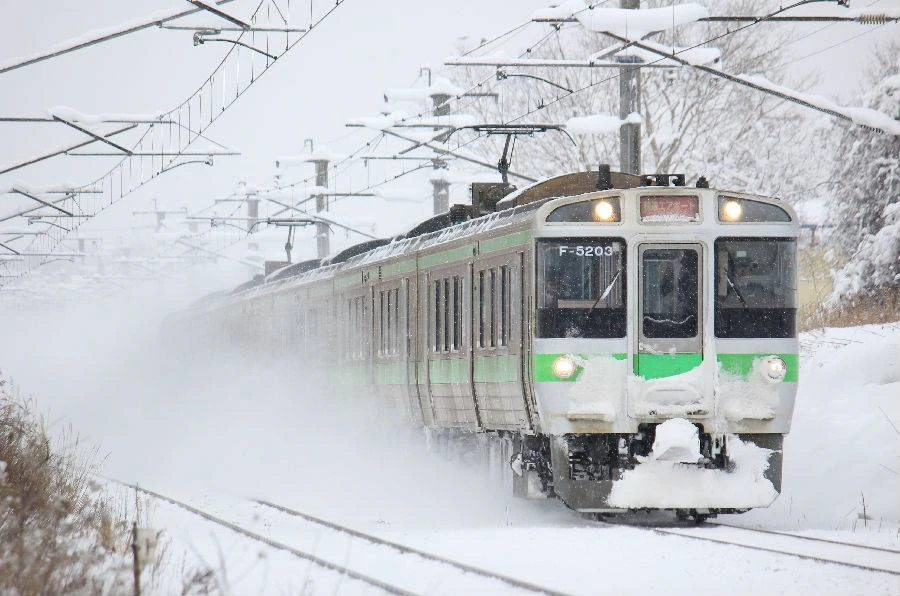 A train moves through a snowy landscape, with snow covering the tracks and surrounding area.
