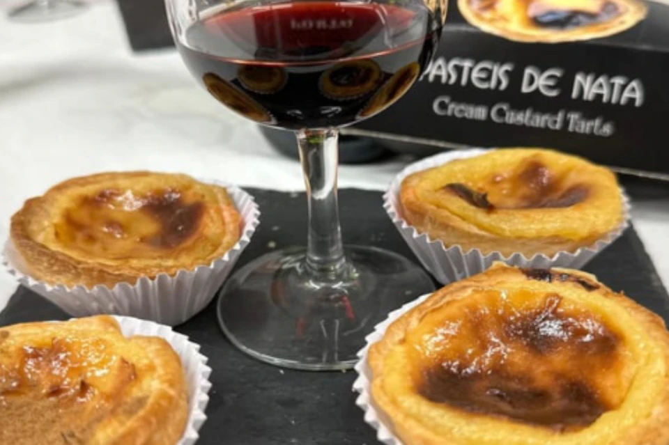 A glass of red wine and four Portuguese custard tarts on a black surface, with part of a box visible in the background.