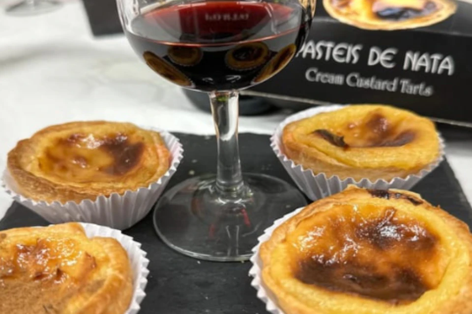 A glass of red wine and four Portuguese custard tarts on a black surface, with part of a box visible in the background.