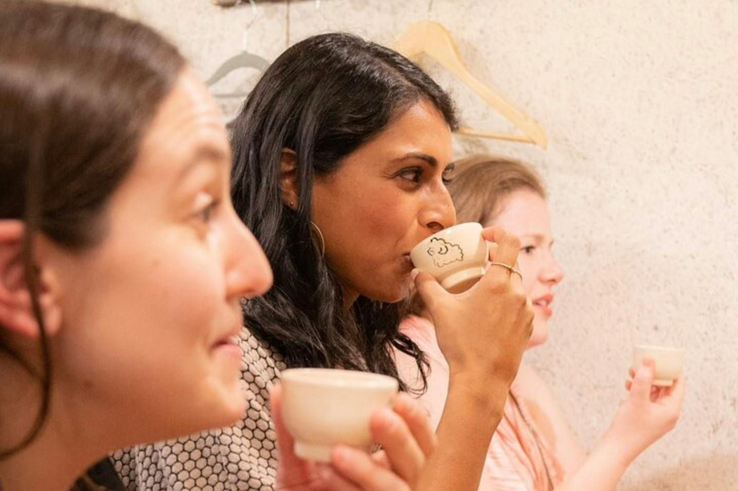 Three women sitting together, drinking from small cups, with a wall and coat hangers visible in the background.
