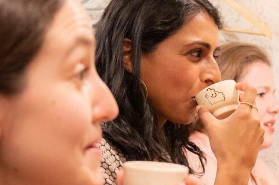 Three people enjoying drinks from small cups, sitting indoors near a wall with coat hangers in the background.