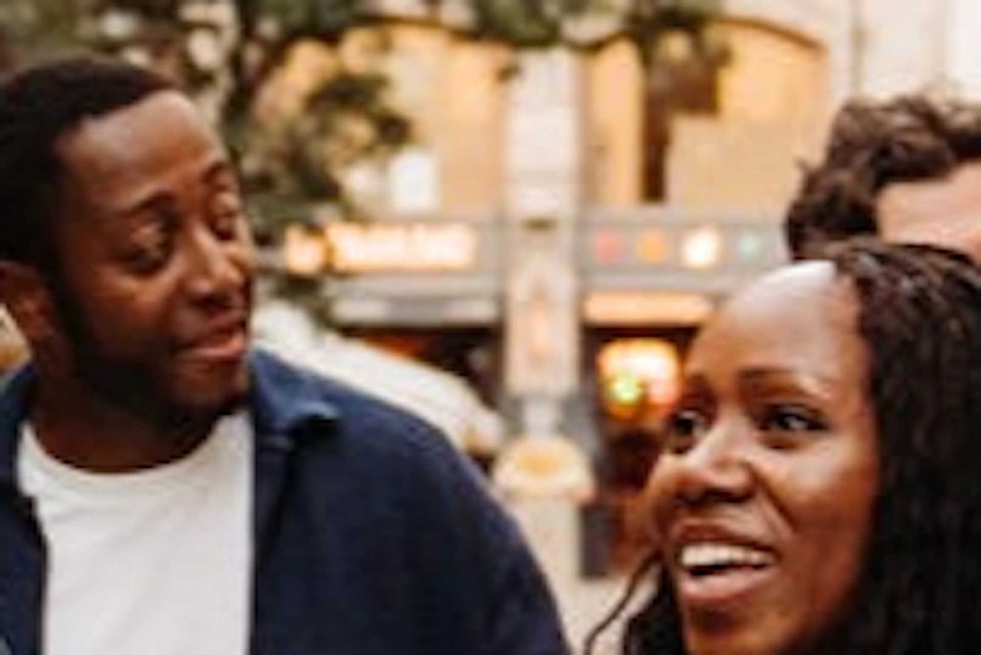 A group of people walking down a lively street with shops, trees, and outdoor cafes in the background.
