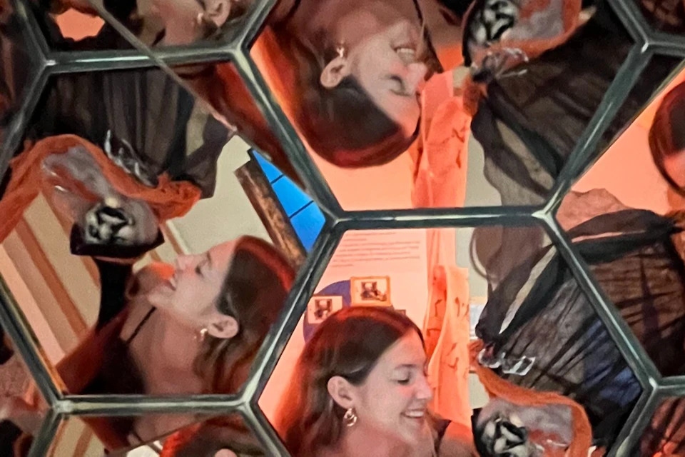 A kaleidoscopic image showing multiple reflections of a smiling woman in a geometric mirrored structure.