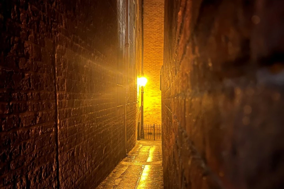 A narrow alleyway illuminated by a warm streetlight, with wet reflective surfaces and brick walls on both sides.