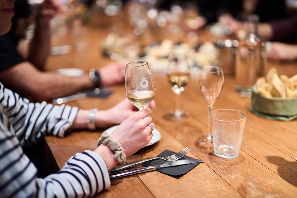 A hand holds a wine glass on a wooden table set with cutlery and glasses, with other people seated in the background.