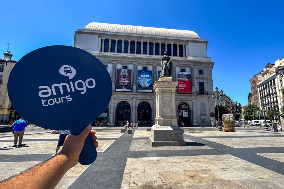 A hand holds a sign labeled 'Amigo Tours' in front of a historic building with a statue in a sunny plaza.