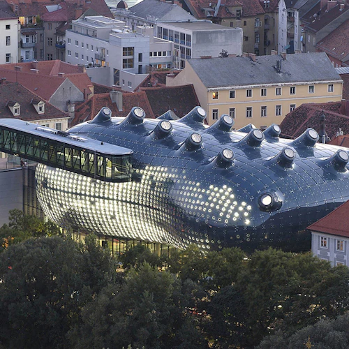 En modern, glödande byggnad med flera runda fönster och utskjutande delar, omgiven av traditionella byggnader och frodiga träd.