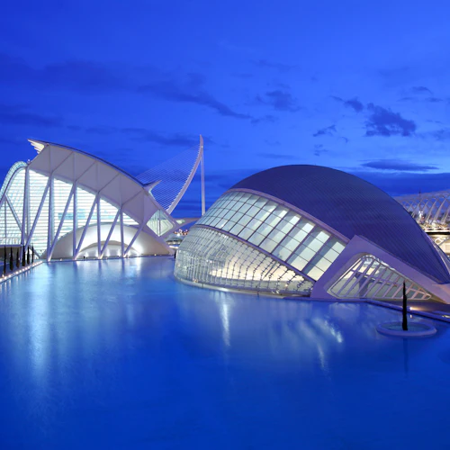 Moderna futuristiska byggnader med vita strukturer och stora glasfönster, omgivna av en reflekterande pool under en skymningshimmel.