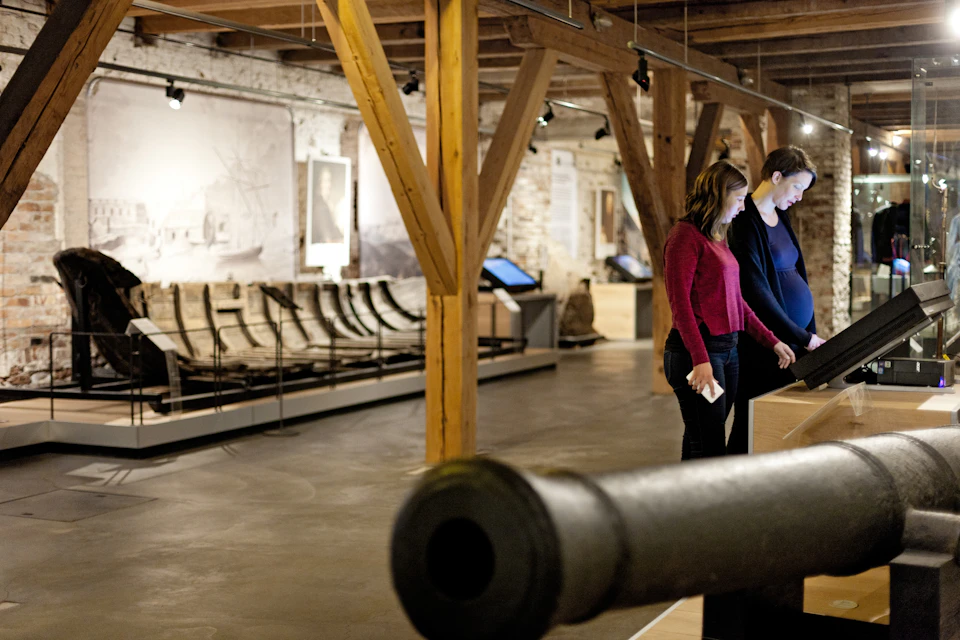 Two people interact with exhibits in a museum with wooden beams, historical artifacts, and black-and-white photos on the walls.