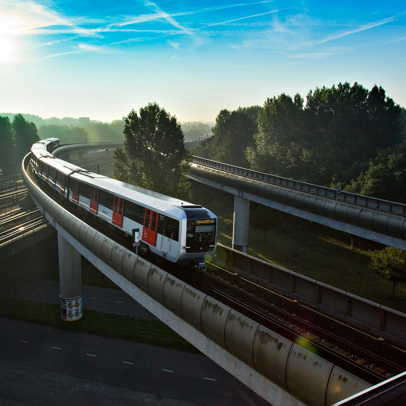 Um comboio viaja numa via elevada através de uma área arborizada sob um céu azul claro.