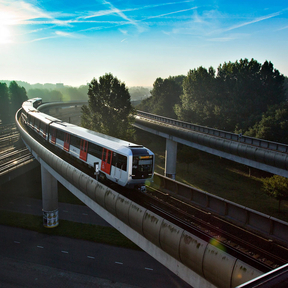 A train travels on an elevated track through a tree-lined area under a clear blue sky.