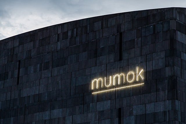 A dark, modern building facade with the illuminated word "mumok" in neon lights against a cloudy sky.
