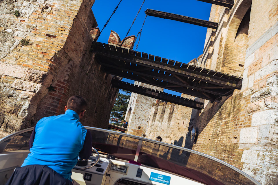 A person navigates a motorboat under a stone bridge with chains, surrounded by ancient brick walls and a clear blue sky above.