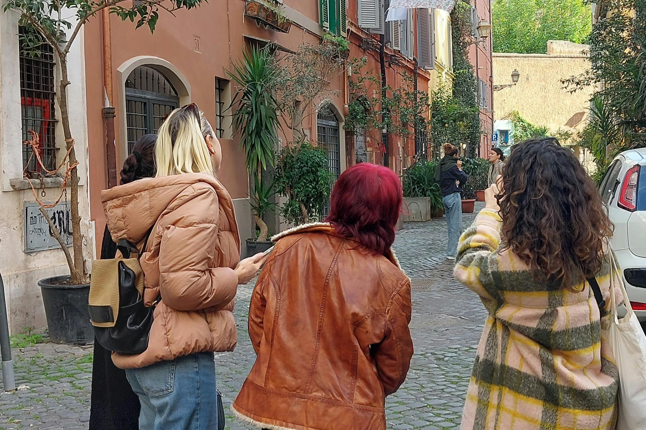 A group of people stand on a cobblestone street lined with colorful buildings and lush plants, engaging in conversation.