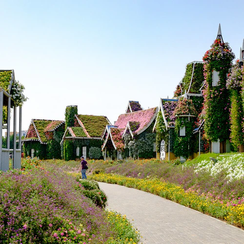 A flower-lined path leads to houses fully covered in greenery and flowers under a clear sky; a person stands in the distance.