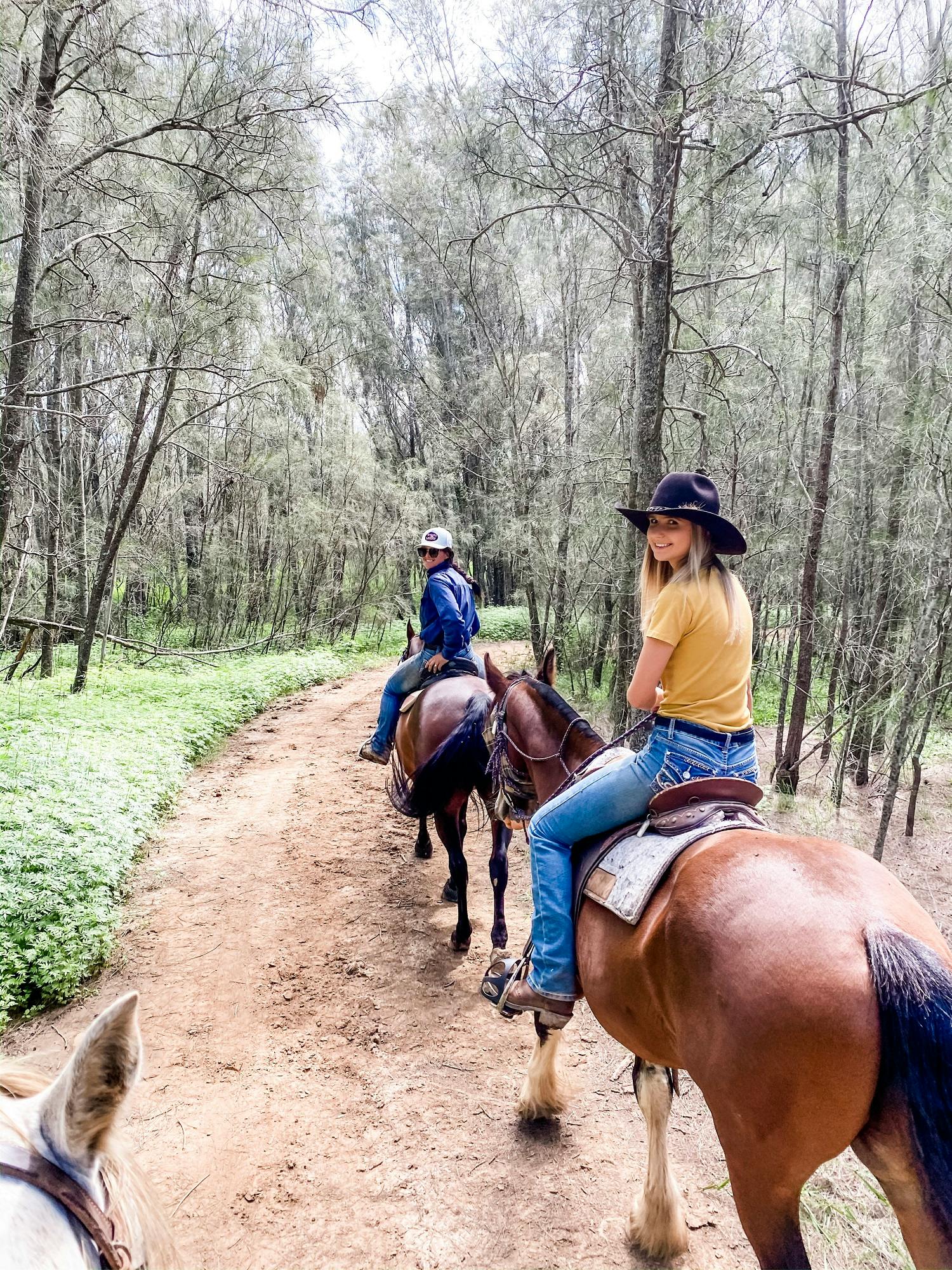 Two people riding horses on a dirt path in a forested area, with trees and greenery surrounding them.