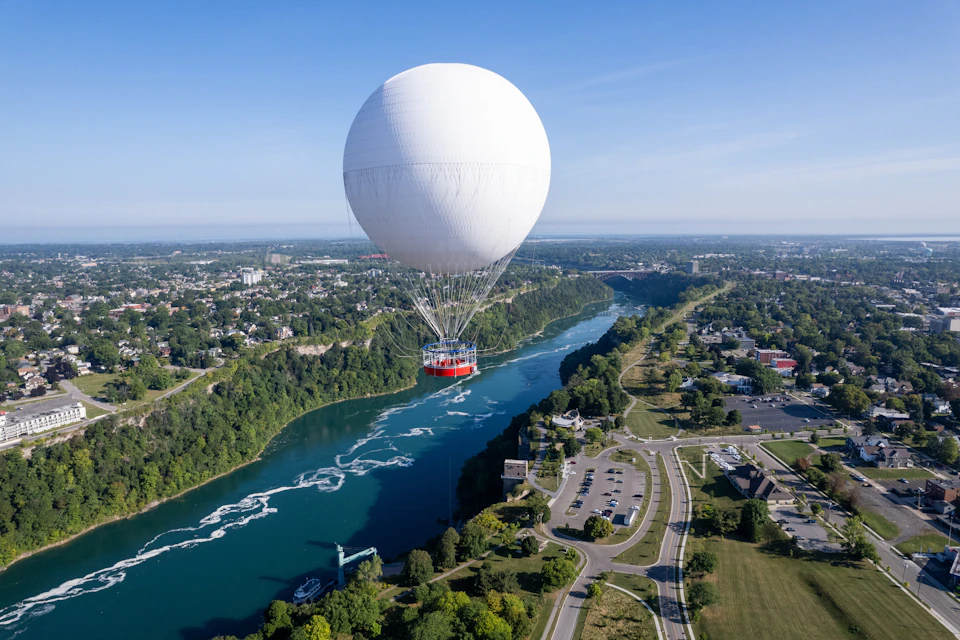 A large white tethered hot air balloon floating above a river, with a surrounding landscape of trees, roads, and buildings.