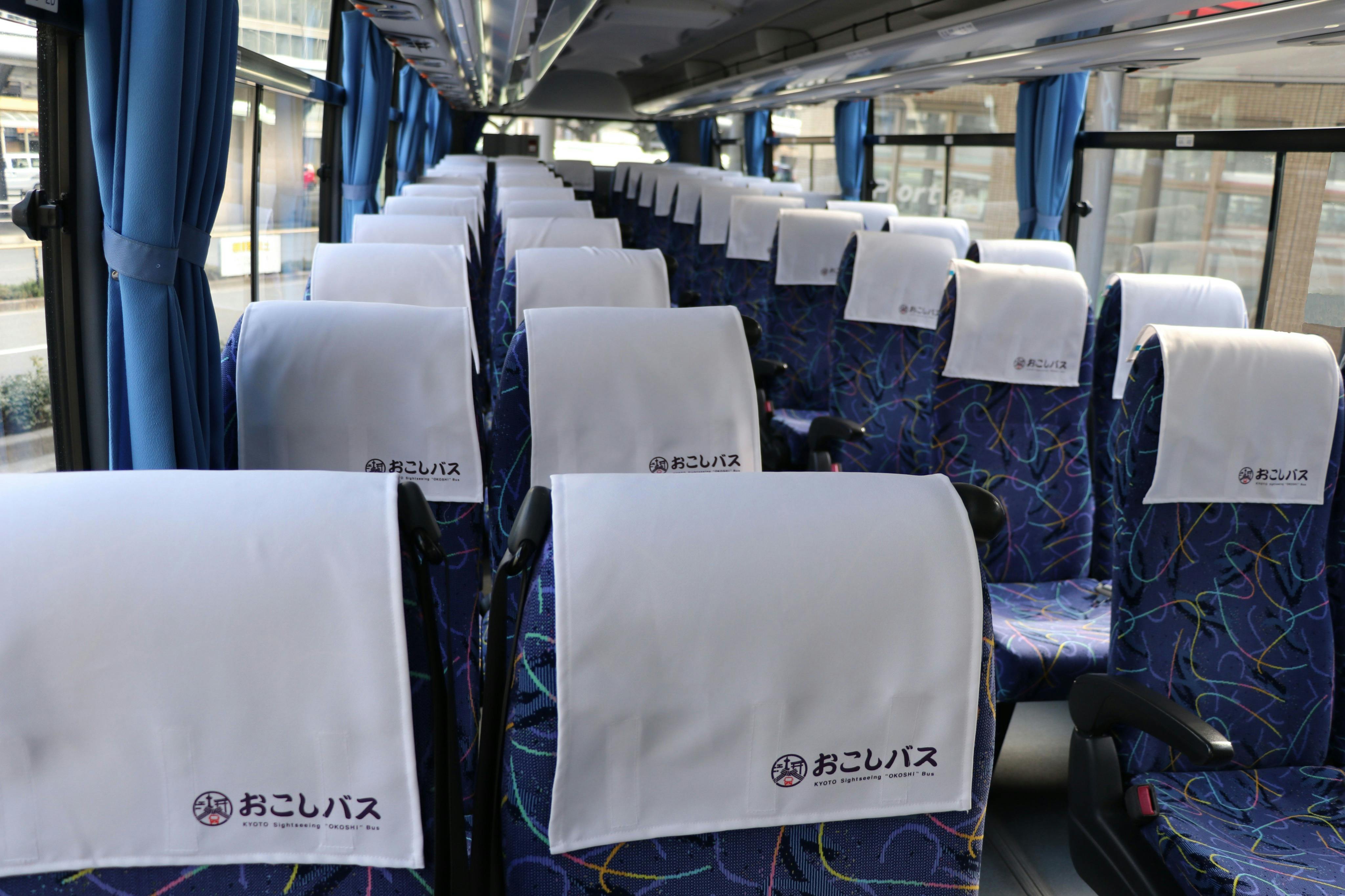 Εσωτερικό ενός καθαρού, άδειου λεωφορείου με σειρές καθισμάτων με μπλε μοτίβα, το καθένα με λευκό κάλυμμα προσκεφάλου με κείμενο στα ιαπωνικά.