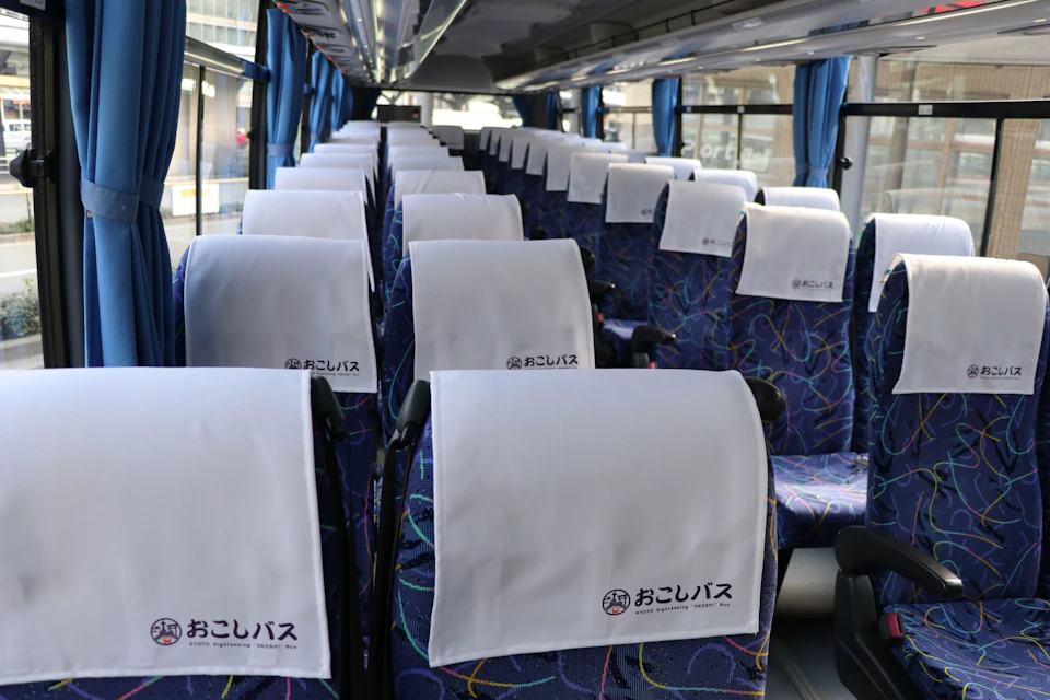 Interior of a clean, empty bus with rows of blue-patterned seats, each with a white headrest cover featuring text in Japanese.