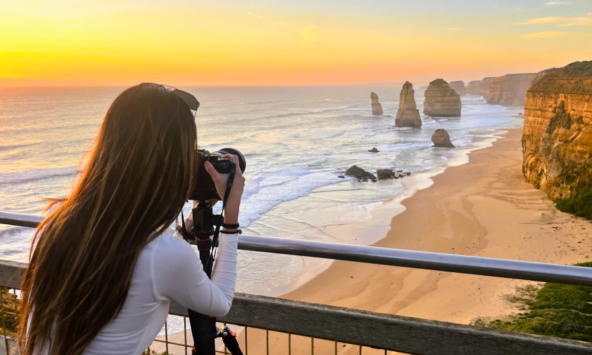 Person with a camera on a tripod photographing the ocean at sunset, with rock formations and a sandy beach in the background.