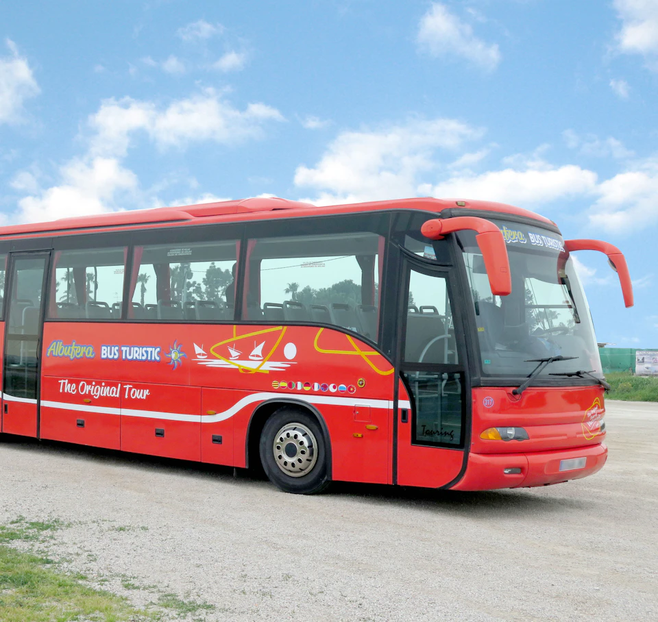 A red tour bus parked on a gravel area under a partly cloudy blue sky, displaying "The Original Tour" and various logos on the side.
