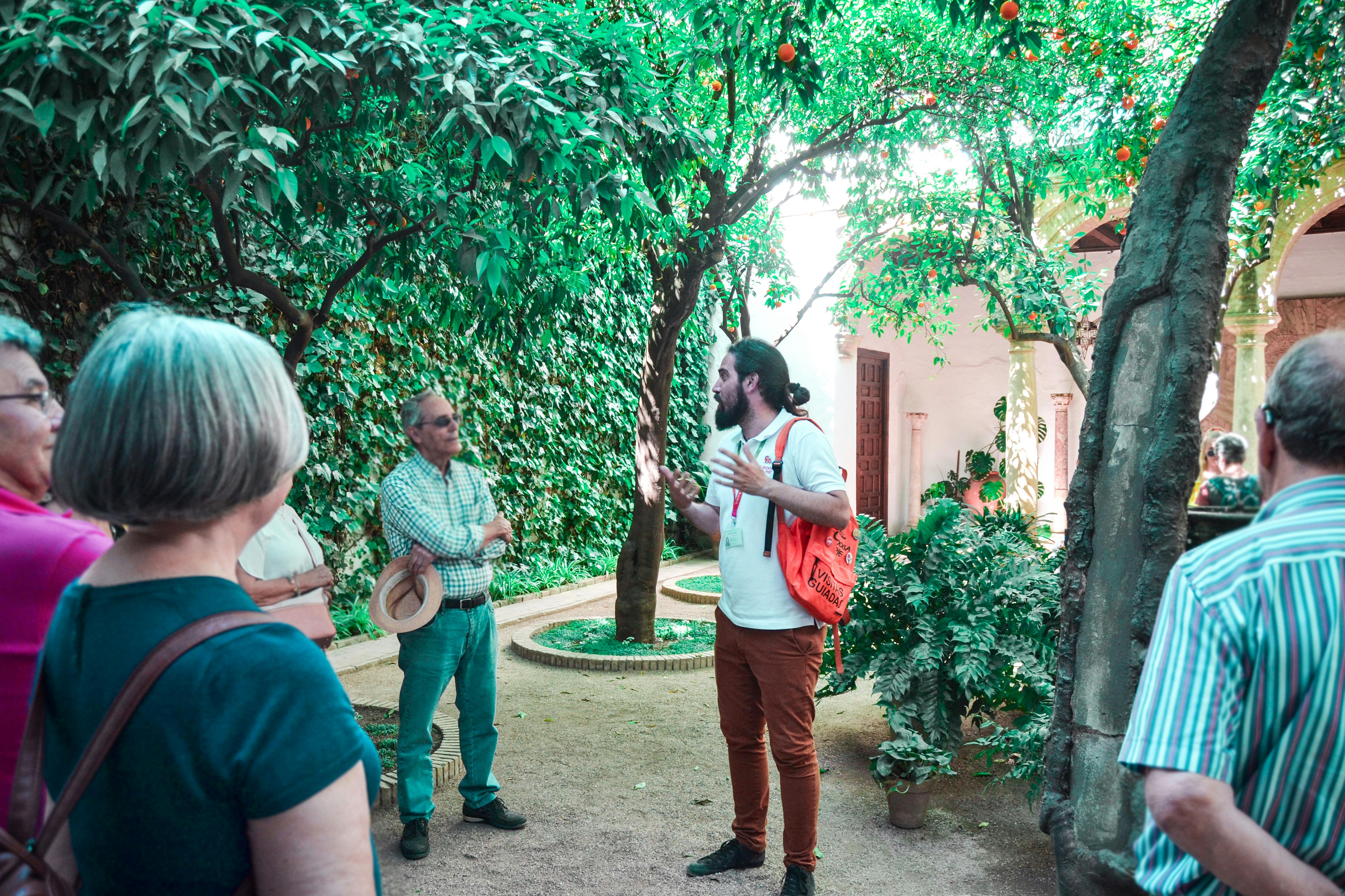 一位背着橙色背包的导游在郁郁葱葱的花园里与一小群人交谈，花园里有树木和常春藤覆盖的墙壁。