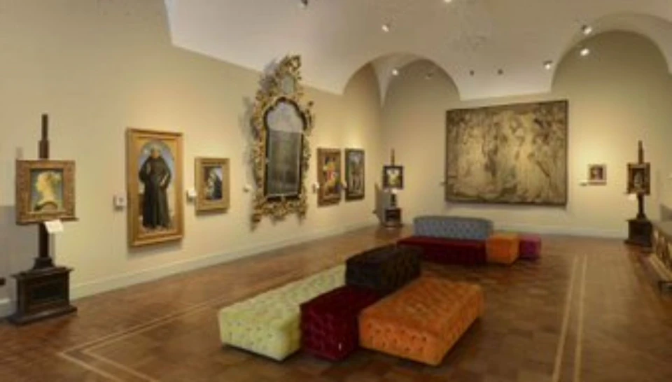 A gallery room with various framed art pieces on the walls and colorful, tufted ottomans arranged in the center.
