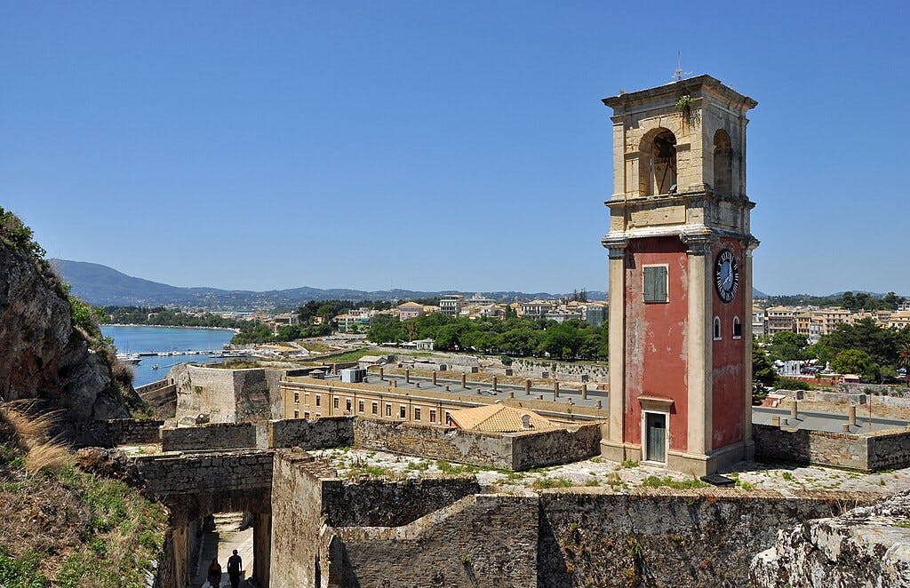 A red clock tower in a historical fortress overlooking a coastal town and blue sea, with mountains in the background.
