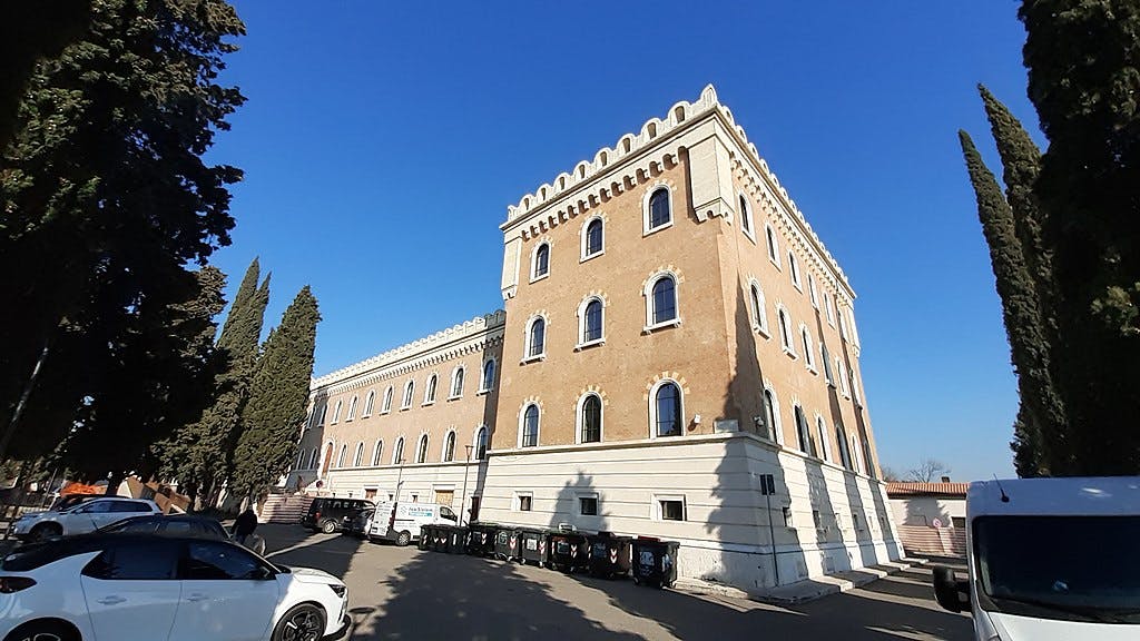 Un alto edificio storico con finestre ad arco, affiancato da alti alberi e auto parcheggiate, sotto un cielo azzurro e limpido.