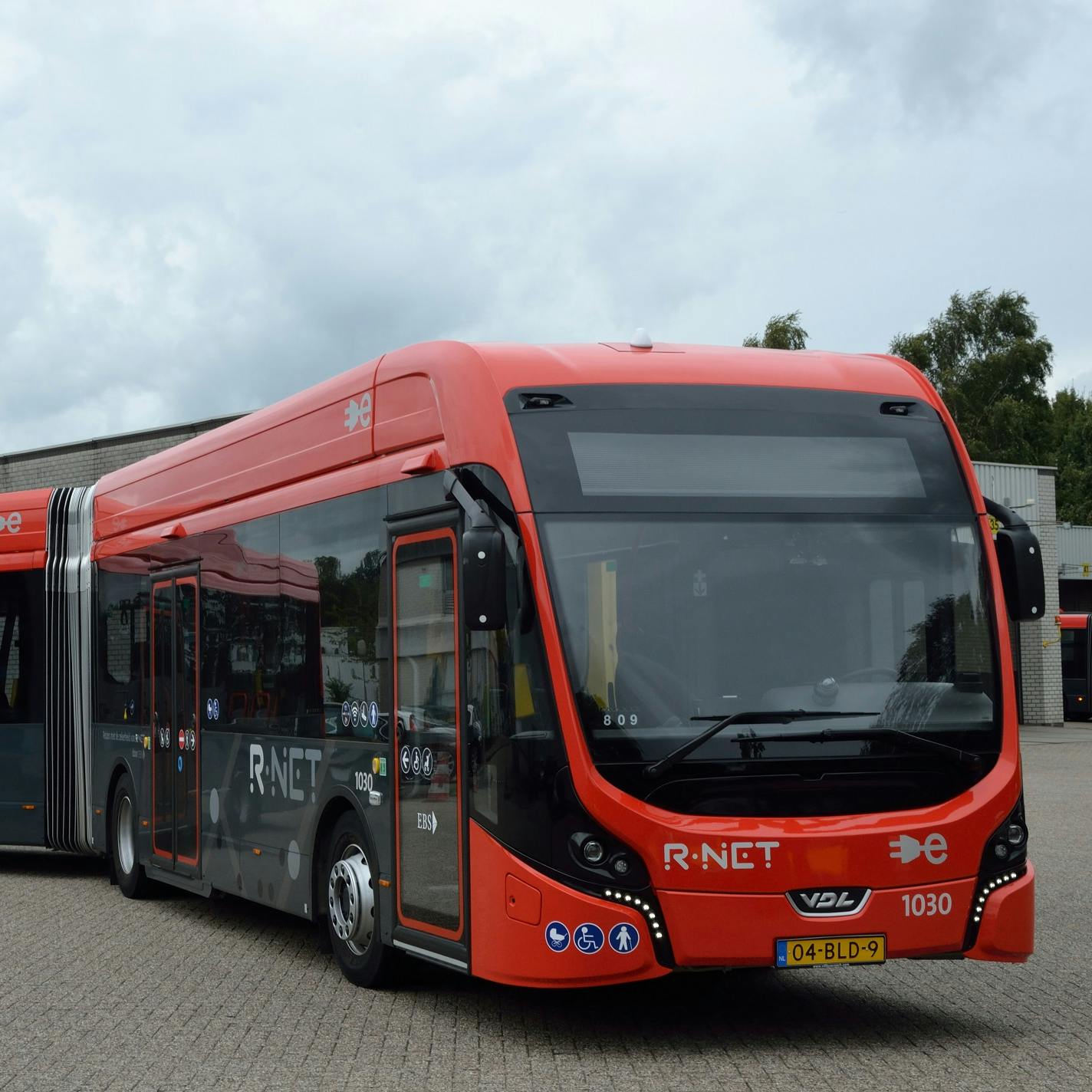A red and black articulated bus with "R-NET" on the side and a Dutch license plate is parked on a paved area.
