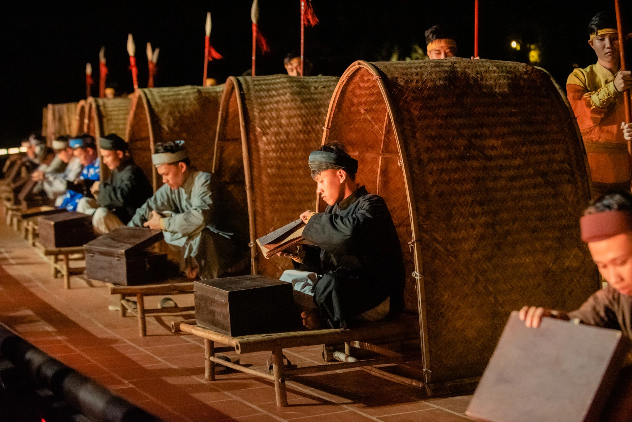 People wearing traditional clothing seated near woven bamboo structures, with a nighttime setting and dim lighting.