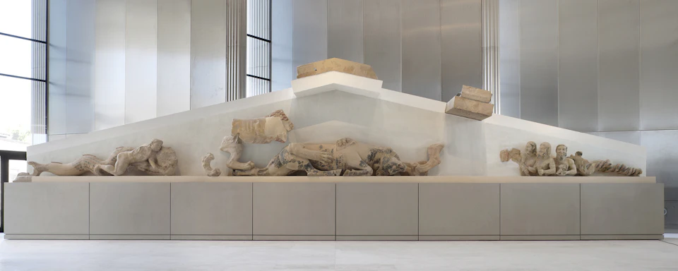 Stone sculptures of reclining and seated human figures are displayed on a raised platform in a modern, minimalist gallery setting.