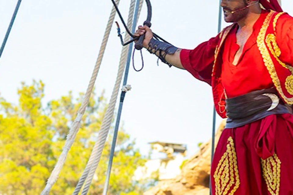 A theatrical scene on a pirate ship with actors in period costumes, including a man in red and a woman in white and green.
